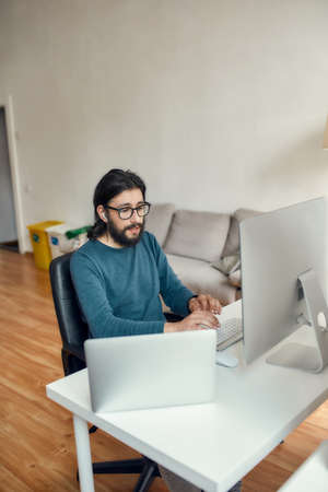 Young focused caucasian bearded man using computer while sitting at his workplace and working from home. Freelance, web developer working onlineの写真素材