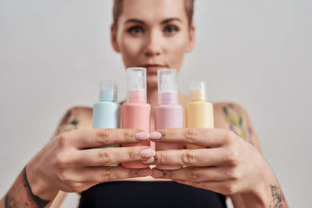 Portrait of tattooed woman with short hair holding plastic bottles with cosmetic, hair and skin care products, isolated over grey backgroundの写真素材