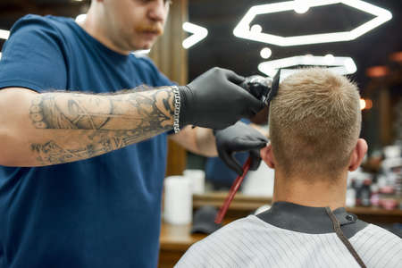 Professional tattooed barber working with hair clipper. Man visiting barbershop and getting new modern haircut. Focus on a clients headの写真素材