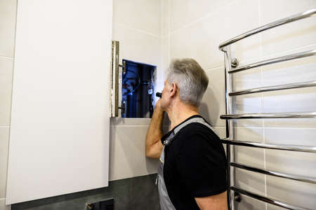 Aged plumber, repairman in uniform examining, inspecting pipes using flashlight in the bathroom....