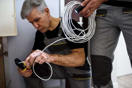 Fix it. Cropped shot of aged electrician, repairman in uniform working, examining cable, checking voltage in fuse box using multimeter while his colleague holding cableの写真素材