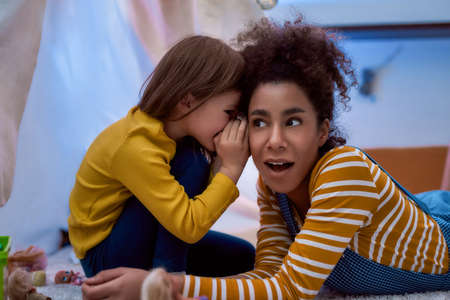 Listening is a key. African american woman baby sitter entertaining caucasian cute little girl. They are gossiping and telling secrets sitting in kids room. Leisure activities, babysitting conceptの写真素材