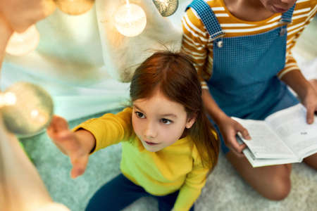Right environment for child. African american woman baby sitter entertaining caucasian cute little girl, reading a book to her. Kid enjoying sitting in wigwam, tentの写真素材