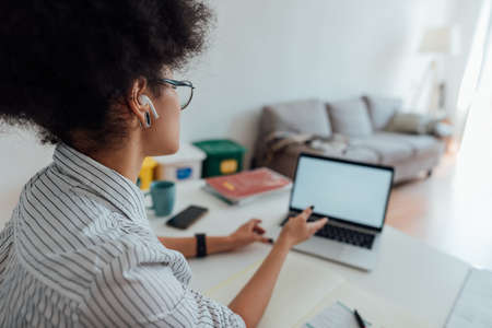 Rear view of afro american woman wearing wireless headphones looking at webcam and teaching English online while working from homeの写真素材