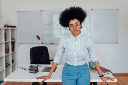 Portrait of young professional afro american woman in casual clothes looking at camera while teaching foreign language online from home. Whiteboard on the backgroundの写真素材