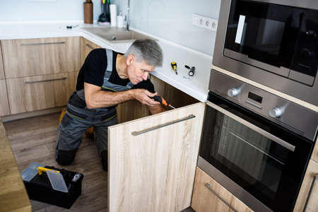 Five star service. Aged repairman in uniform working, fixing kitchen cabinet using screwdriver. Repair service conceptの写真素材