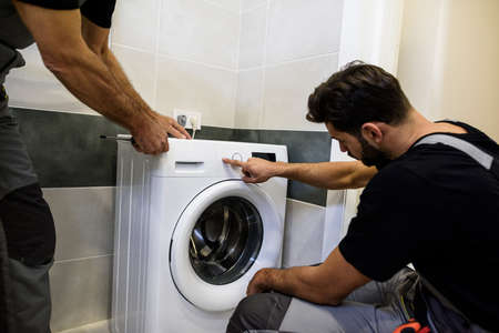 Two repairmen, workers in uniform working, reading washing symboles on washing mashine before fixing it, holding screwdriver in the bathroom. Repair service conceptの写真素材