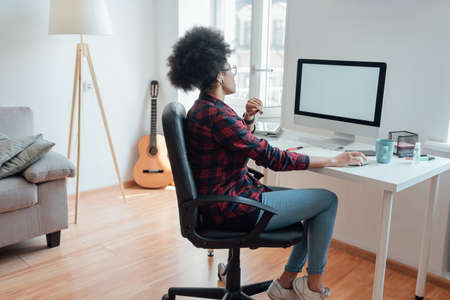 Comfortable workplace. Side view of afro american woman looking at computer screen while working or learning online from homeの写真素材