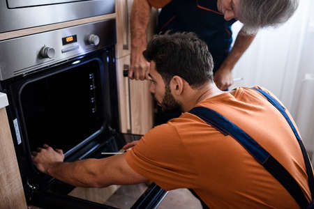 Close up shot of two repairmen, workers in uniform examining oven using screwdriver and flashlight indoors. Repair service conceptの写真素材