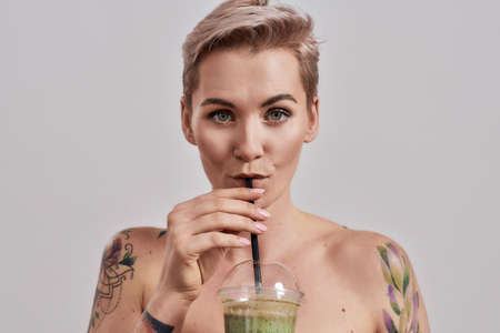 Attractive tattooed woman with pierced nose and short hair looking at camera, while drinking green detox smoothie isolated over grey backgroundの写真素材