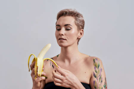 Attractive tattooed woman with pierced nose and short hair looking at banana while peeling it...