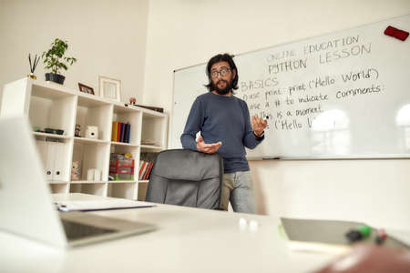 Online classroom. Young caucasian man teacher in glasses standing near whiteboard and teaching Python, giving online class at homeの写真素材