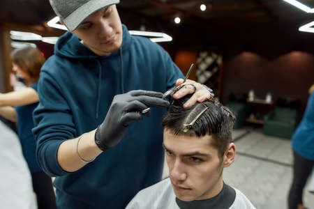 Professional male barber making new trendy haircut for a young handsome guy sitting in barbershop...