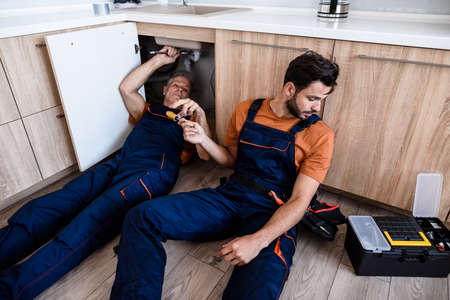 Young repairman, worker in uniform sitting on the floor, bringing screwdriver while his experienced colleague fixing sink pipe indoors. Repair service conceptの写真素材