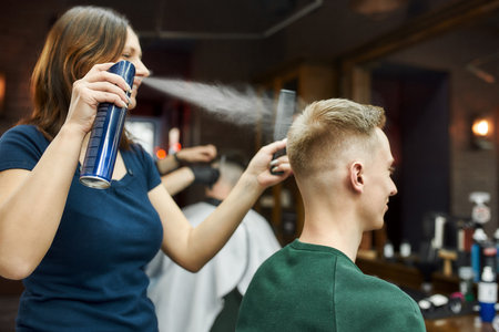 Side view of a professional barber girl fixing new modern haircut with hairspray of a young guy sitting in barber shop chairの写真素材