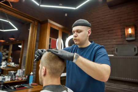 Working process. Professional barber serving client, making trendy haircut for young caucasian man visiting modern barbershopの写真素材