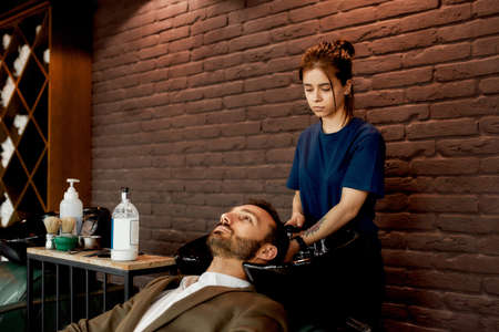 Young professional barber girl, hairdresser washing hair of a handsome bearded man leaning on the...
