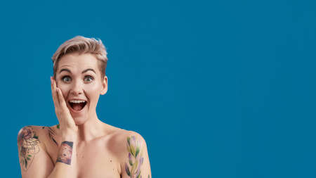 Portrait of a young attractive half naked tattooed woman with perfect skin looking excited, holding hand on her cheek isolated over blue backgroundの写真素材
