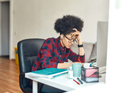 Exhausted afro american woman studying online at home. Sitting at her comfortable workplace, using computer and making some notesの写真素材