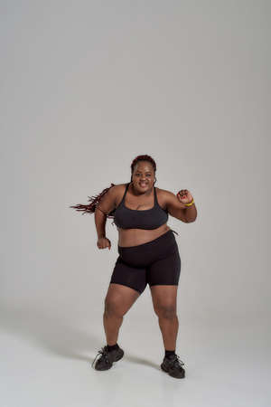 Full length shot of plump, plus size african american woman in sportswear having fun, dancing in studio over grey backgroundの写真素材