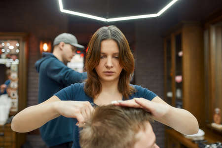 Young professional barber girl making haircut for client sitting in a barbershop chair. Focus on a womanの写真素材