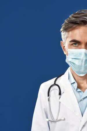 Half face portrait of mature male doctor wearing medical protective mask, looking at camera while standing against blue navy backgroundの写真素材