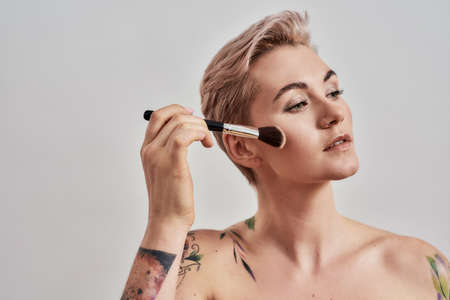 Gentle Touch. Portrait of beautiful tattooed woman with pierced nose and short hair applying makeup, blush with a brush isolated on grey backgroundの写真素材