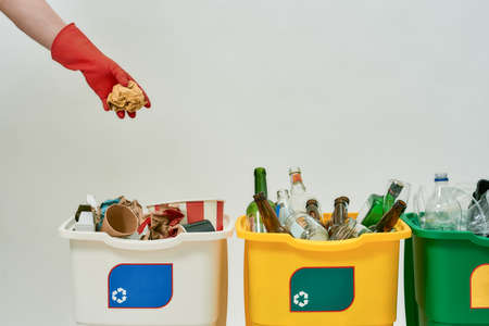 Hand in red glove throwing paper into one of three colorful green, yellow and white recycle garbage bins full of rubbish isolated on white background. Waste separation conceptの写真素材