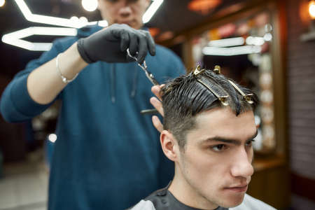Handsome young guy visiting barbershop, getting new modern haircut. Professional barber holding scissors and making haircut for his clientの写真素材