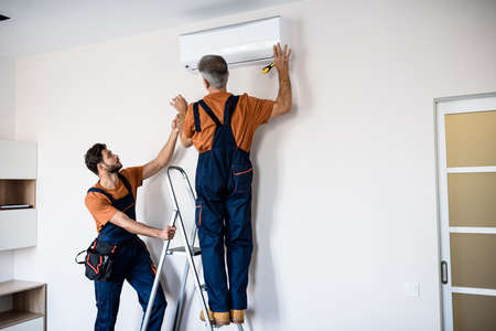 Two workers in uniform, air conditioning masters using ladder while installing a new air conditioner in the apartment. Construction, maintenance and repair conceptの写真素材