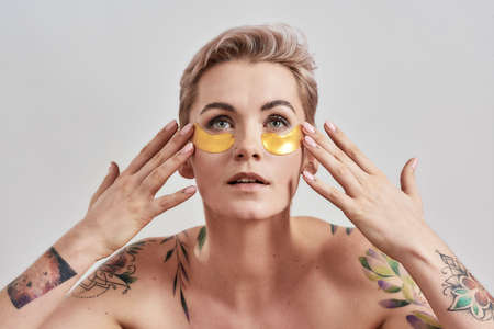 Beauty portrait of half naked tattooed woman with short hair looking up, caring for her face, wearing under eye patches for dark circles isolated over light backgroundの写真素材