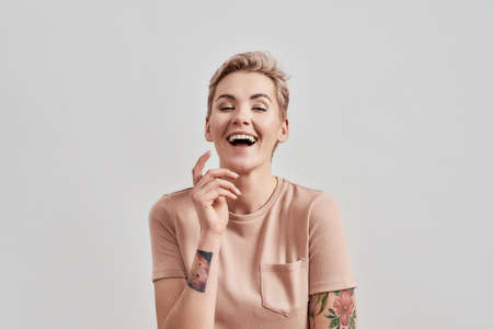 Portrait of a young attractive tattooed woman with pierced nose and short hair in beige t shirt laughing at camera, posing with hand near face isolated over light backgroundの写真素材