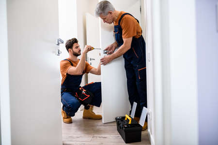 Full length shot of two locksmith, repairmen, workers in uniform installing, working with house door lock using screwdriver. Repair, door lock service concept