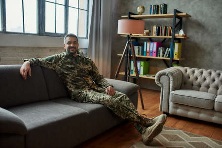 Relive. Full length shot of middle aged military man smiling at camera, sitting on the couch during therapy session. Soldier suffering from depression, psychological trauma. PTSD conceptの写真素材