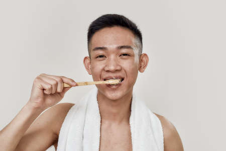 Clean teeth. Portrait of shirtless young asian man with towel around his neck smiling at camera while brushing his teeth isolated over white background. Beauty, skincare, morning routineの写真素材
