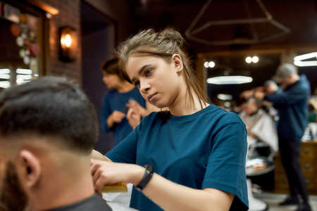 Serving client in Barbershop. Professional barber girl, female hairdresser making modern haircut for a man sitting in barber shop chairの写真素材