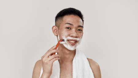 Shave everyday. Portrait of shirtless young asian man with towel around his neck shaving his face, looking at camera isolated over white background. Beauty, skincare, morning routine conceptの写真素材