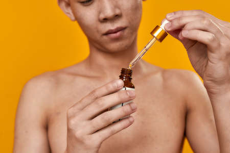 Cropped portrait of young asian man holding gel serum with glass pipette isolated over yellow background. Beauty, skincare routine conceptの写真素材