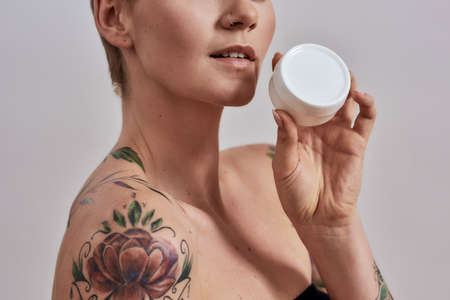 Cropped portrait of beautiful tattooed woman with pierced nose and short hair holding white plastic...