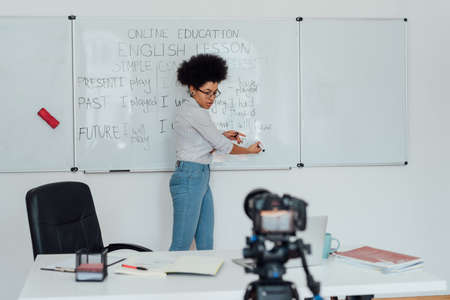 Young afro american woman standing near whiteboard, looking at web camera and explaining rules of English grammar, recording English lesson at homeの写真素材