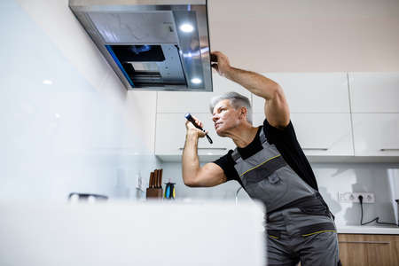 Always quality service. Aged repairman in uniform working, examining broken kitchen extractor fan,...