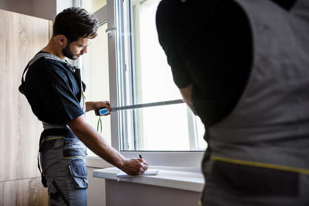 Two professional workers in uniform using tape measure while measuring window and making notes for installing blinds indoors. Construction and maintenance conceptの写真素材