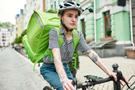 Young delivery woman in helmet with thermo bag or backpack riding a bike along the city, delivering food. Courier, delivery service conceptの写真素材