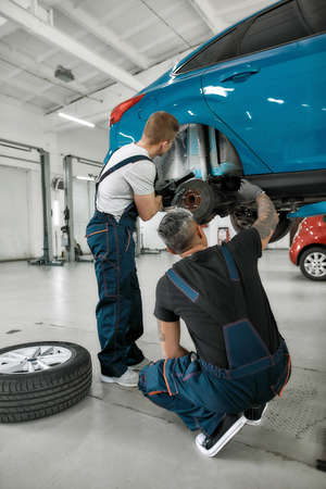 Repairing your vehicle. Full length shot of two male mechanics using torch for examining car wheel brake disc of lifted automobile at auto repair shopの写真素材