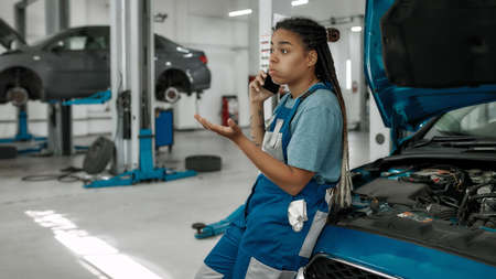 Young african american woman, professional female mechanic talking on phone, leaning on a car with open hood at auto repair shopの写真素材