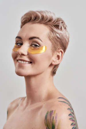 Beauty portrait of half naked tattooed woman with short hair smiling, caring for her face, wearing...
