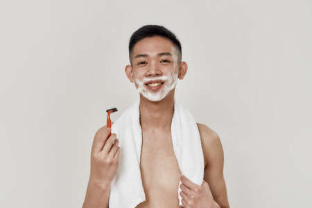 Morning routine. Portrait of shirtless young asian man with towel around his neck looking at camera while shaving his face isolated over white background. Beauty, skincare, morning routine conceptの写真素材