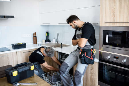 Aged repairman in uniform fixing dishwasher in the kitchen, while his young colleague standing near...
