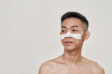 Deep cleansing. Portrait of shirtless young asian man with mask applied on his face looking away isolated over white background. Beauty, skincare routineの写真素材