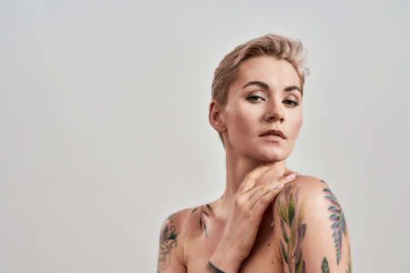 Beauty portrait of a young attractive half naked tattooed woman with perfect skin looking at camera, holding hand near her face isolated over grey backgroundの写真素材